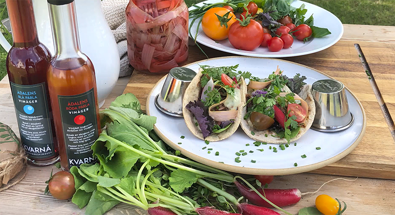 Två fyllda taccobröd ligger på ett fat, på ett bord med grönsaker och vinägerflaskor. Foto.